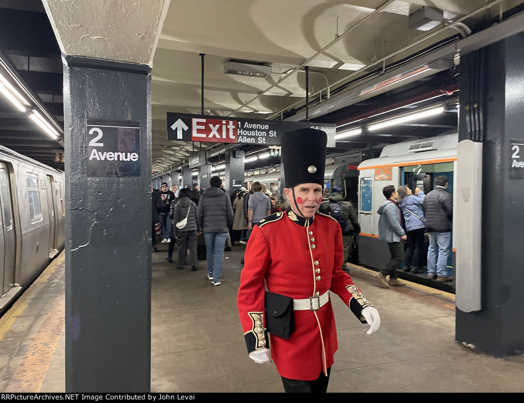 A man wearing a British soldier outfit at 2nd Ave Station walking on the platform between the R179 consist F train and the Arnine consist making up the Holiday Train